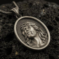 Load image into Gallery viewer, Bronze Jesus Christ pendant with crown of thorns and detailed sacred portrait displayed on dark volcanic rock in a close-up luxury jewelry composition
