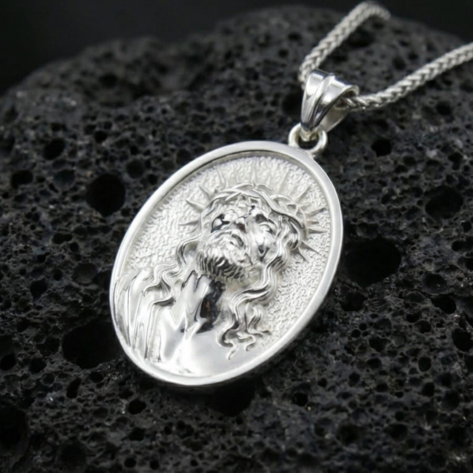 Polished silver Jesus medal with oval frame, crown of thorns, and sculpted devotional face placed on textured volcanic rock in a premium studio product shot