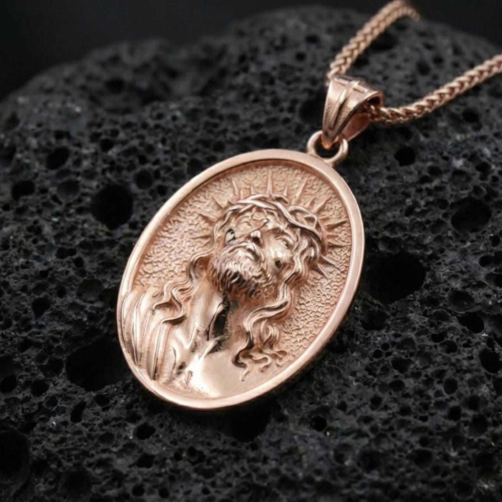 Rose gold plated religious Jesus medal with sacred face relief, crown of thorns, and elongated oval silhouette resting on volcanic rock in an elegant close-up product presentation