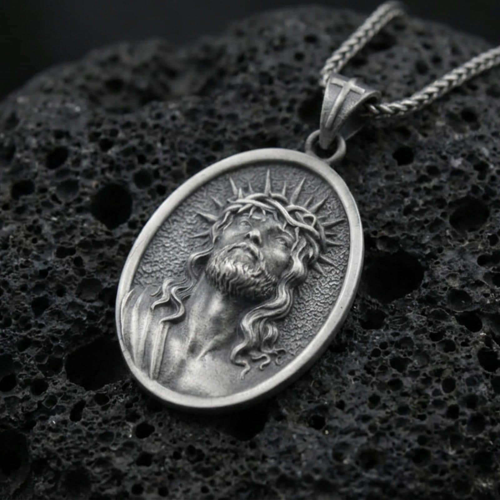 Oxidized Jesus Christ pendant with crown of thorns, radiant halo detail, and sacred portrait displayed on dark volcanic rock in a close-up luxury jewelry composition