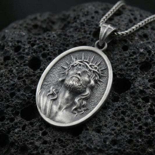 Oxidized Jesus Christ pendant with crown of thorns, radiant halo detail, and sacred portrait displayed on dark volcanic rock in a close-up luxury jewelry composition