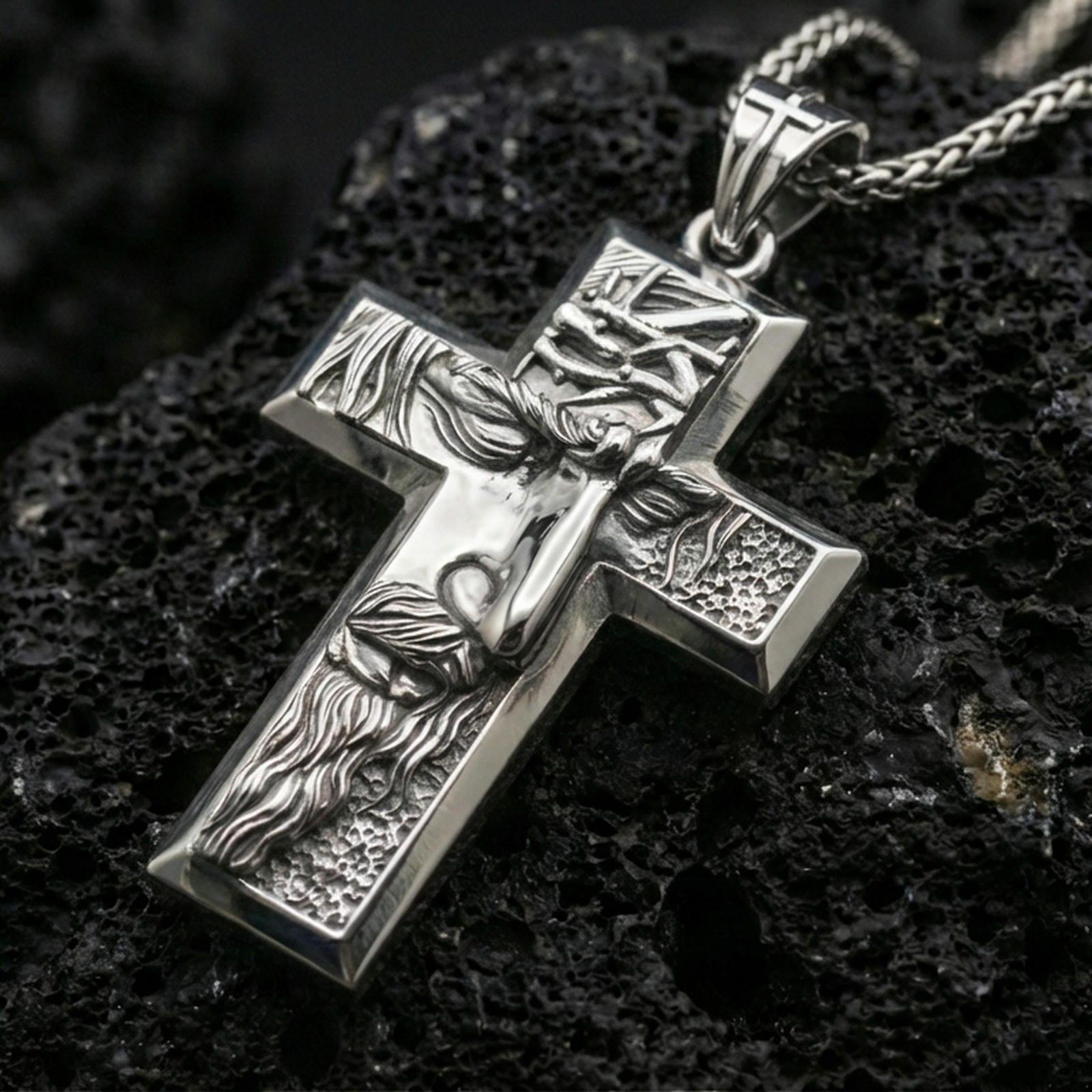 Polished silver Christian cross pendant with detailed Christ figure, bold cross silhouette, and engraved background texture placed on textured volcanic rock in a premium studio product shot