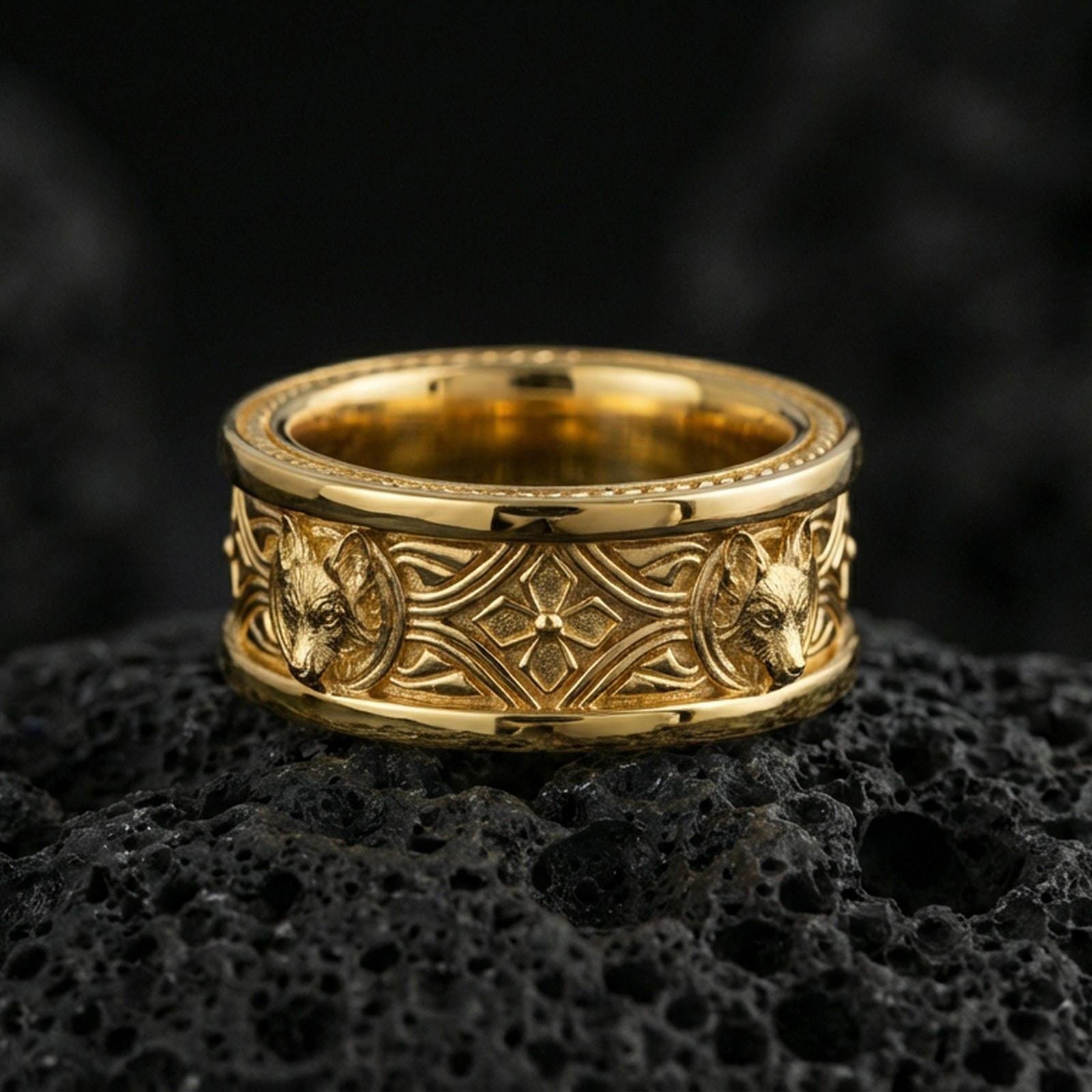Gold plated fox band ring on textured volcanic rock, premium close-up with warm reflective shine across the fox head motifs and engraved ornamental frame.