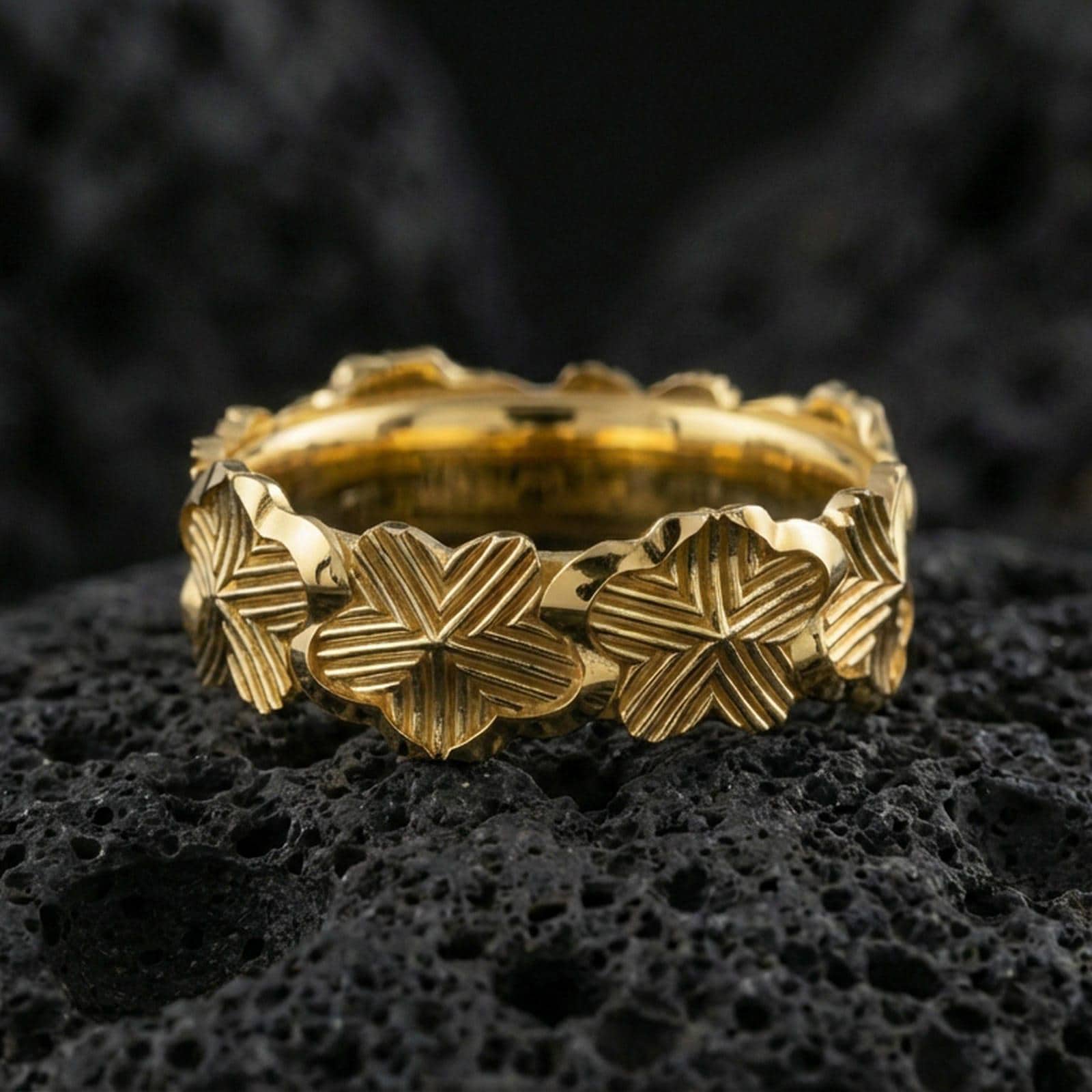 Gold plated floral band ring on textured volcanic rock, premium close-up with warm reflective shine across the lily shapes, sculpted petals, and raised ornamental contours.