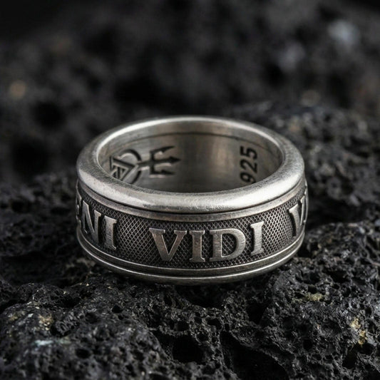 Oxidized Veni Vidi Vici spinner ring on textured volcanic rock, centered close-up highlighting the rotating band, raised Latin lettering, and deep antique shadowed contrast.
