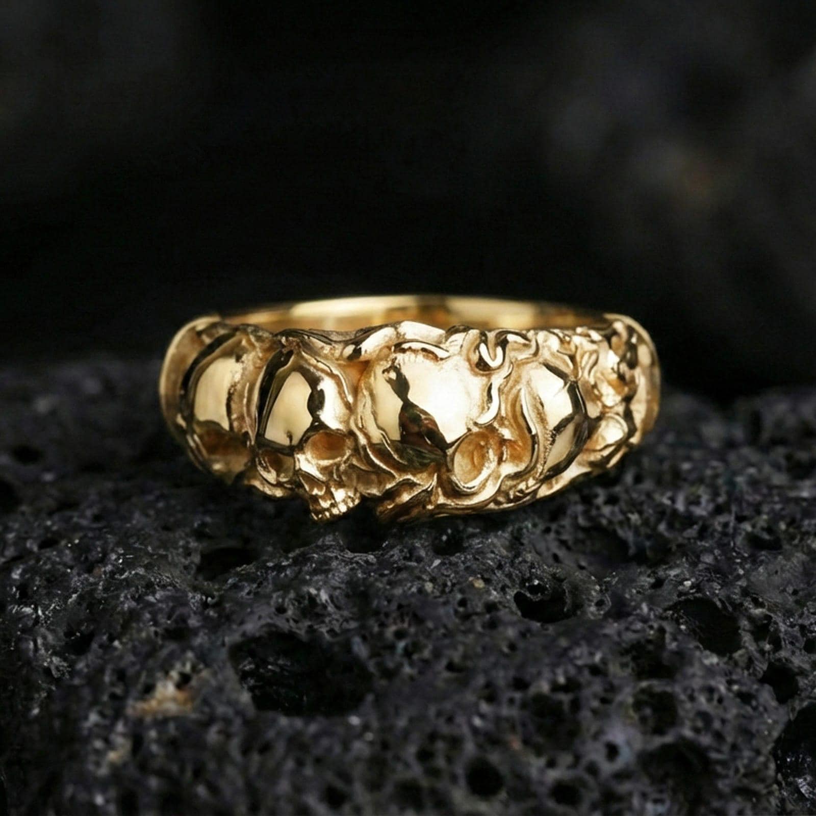 Gold plated skull band ring on textured volcanic rock, premium close-up with warm reflective shine across the skull reliefs and raised sculpted surfaces.