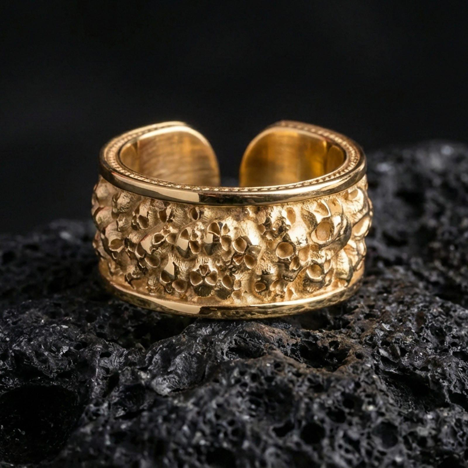 Gold plated skull cuff ring on textured volcanic rock, premium close-up with warm reflective shine across the skull cluster relief and raised sculpted surfaces.