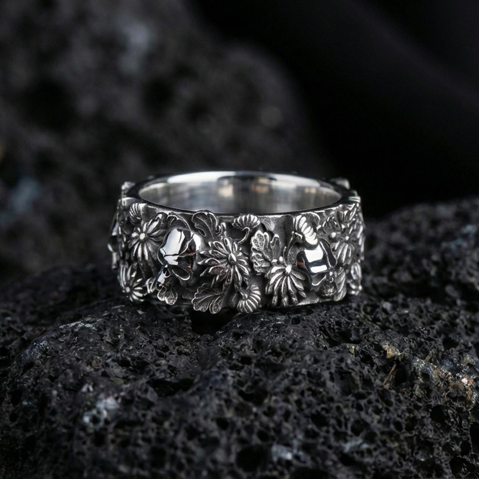 Polished silver skull and floral band ring on textured volcanic rock, crisp macro product photo emphasizing clean edges, sharp petal detailing, and bright reflective highlights across the sculpted band.