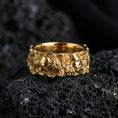 Load image into Gallery viewer, Gold plated skull and floral band ring on textured volcanic rock, premium close-up with warm reflective shine across the skull motifs, floral clusters, and raised ornamental contours.
