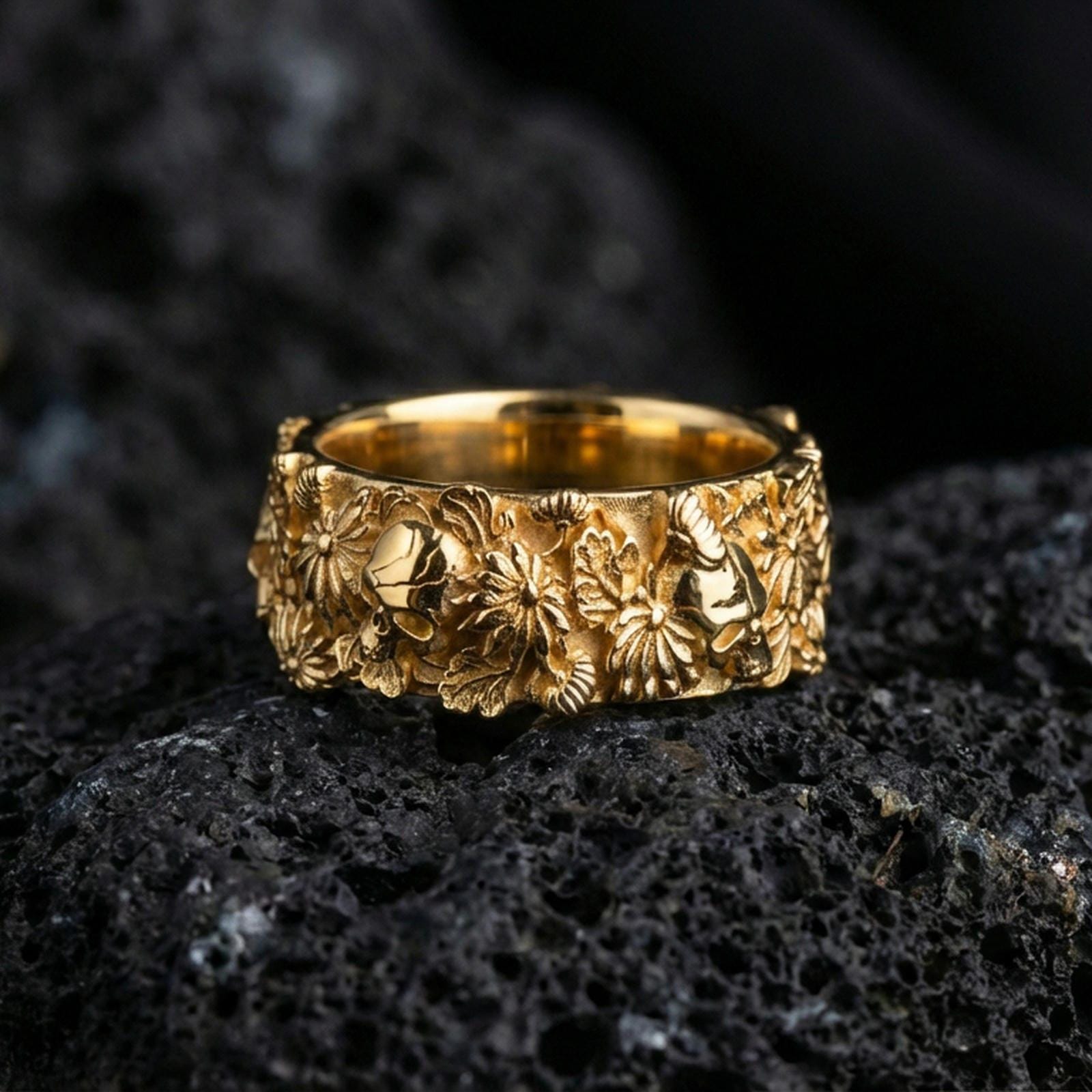 Gold plated skull and floral band ring on textured volcanic rock, premium close-up with warm reflective shine across the skull motifs, floral clusters, and raised ornamental contours.