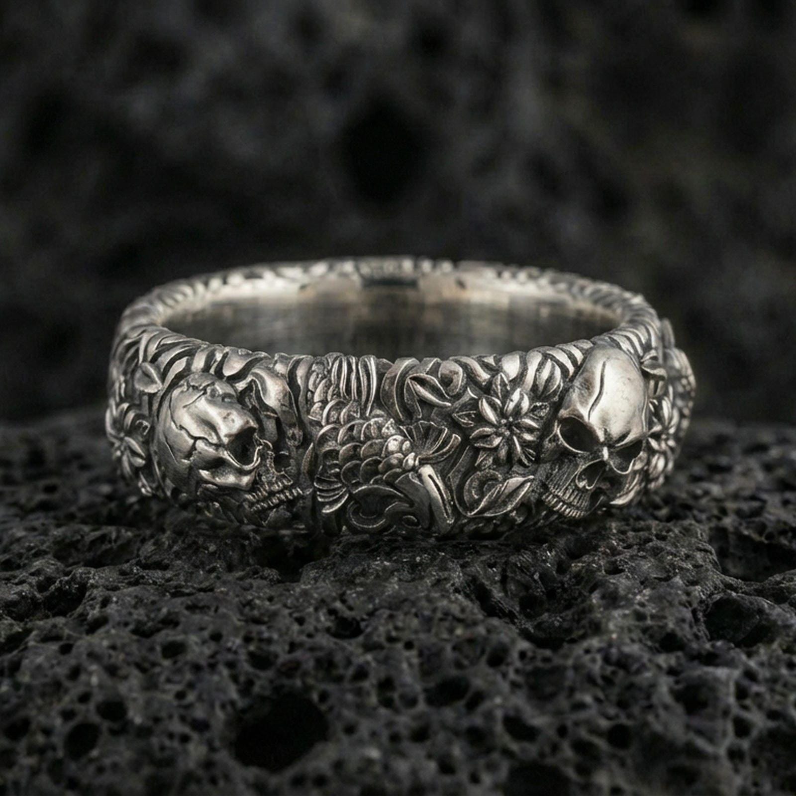 Oxidized skull and koi floral band ring on textured volcanic rock, centered close-up highlighting the raised skull accents, koi scale texture, floral relief, and deep antique shadowed contrast.