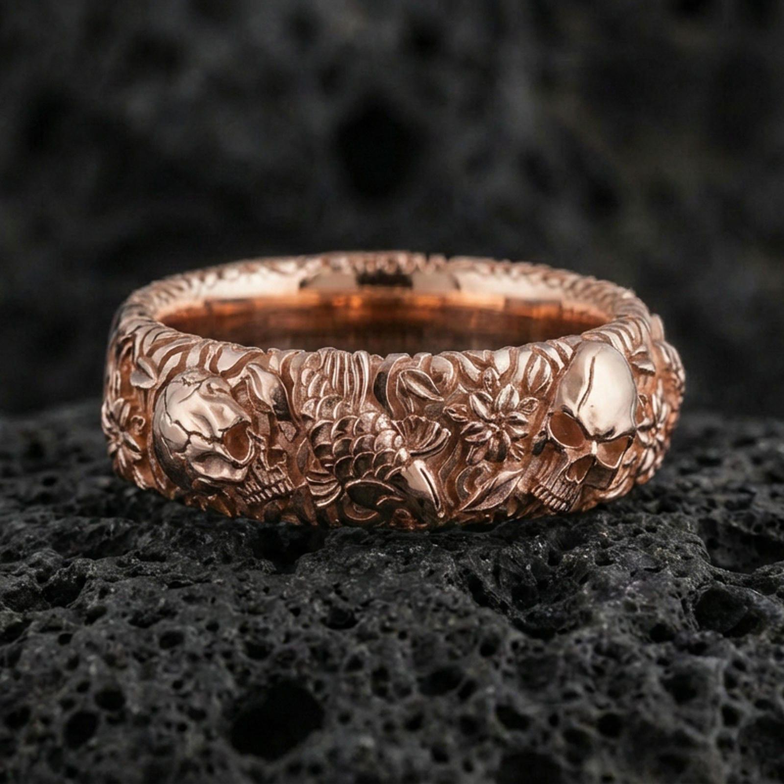Gold plated skull and koi floral band ring on textured volcanic rock, premium close-up with warm reflective shine across the skull motifs, koi detailing, and raised ornamental contours.