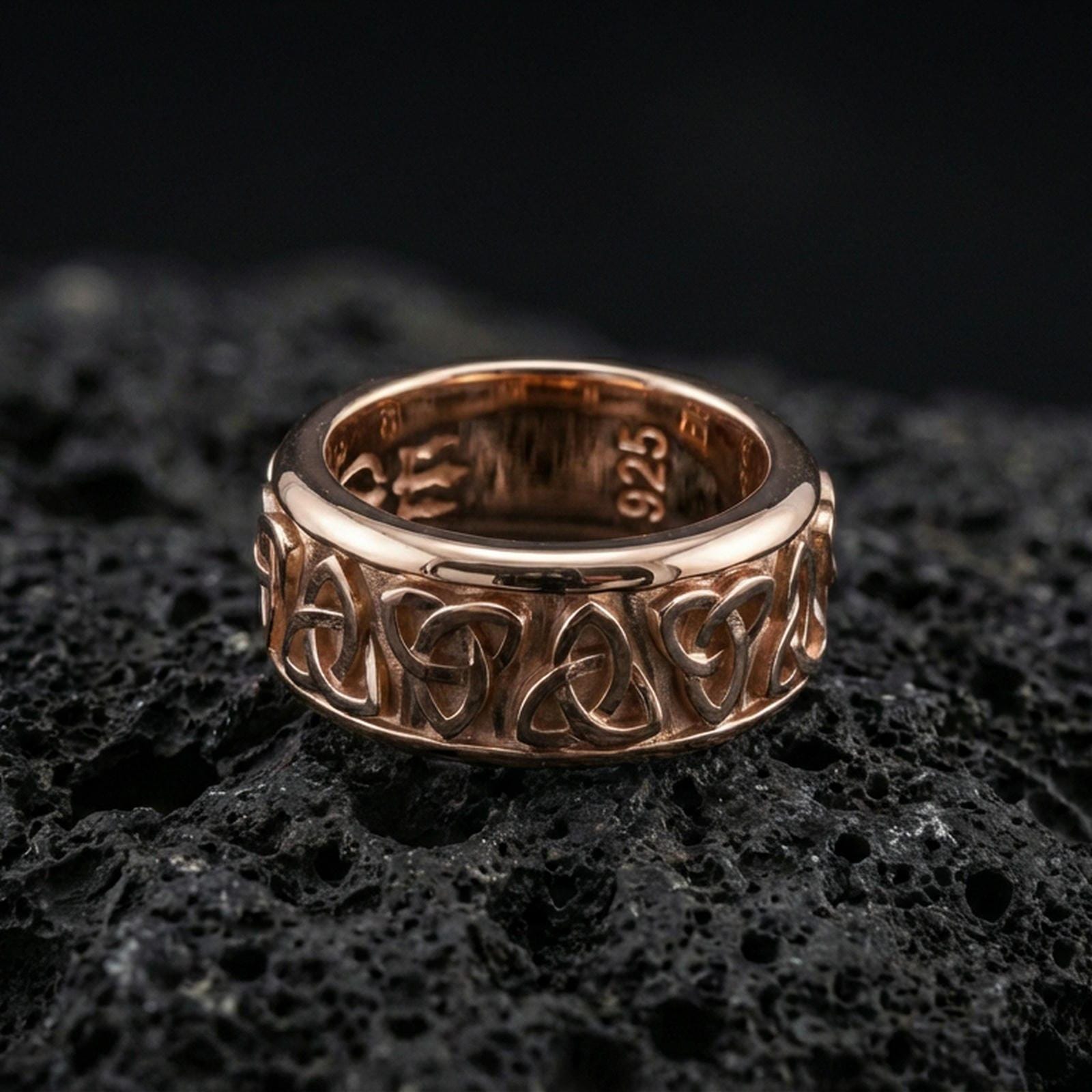 Rose gold plated Celtic triquetra band ring on textured volcanic rock, centered studio-style close-up showcasing soft rosy glow and crisp Celtic knot detailing with a refined finish.