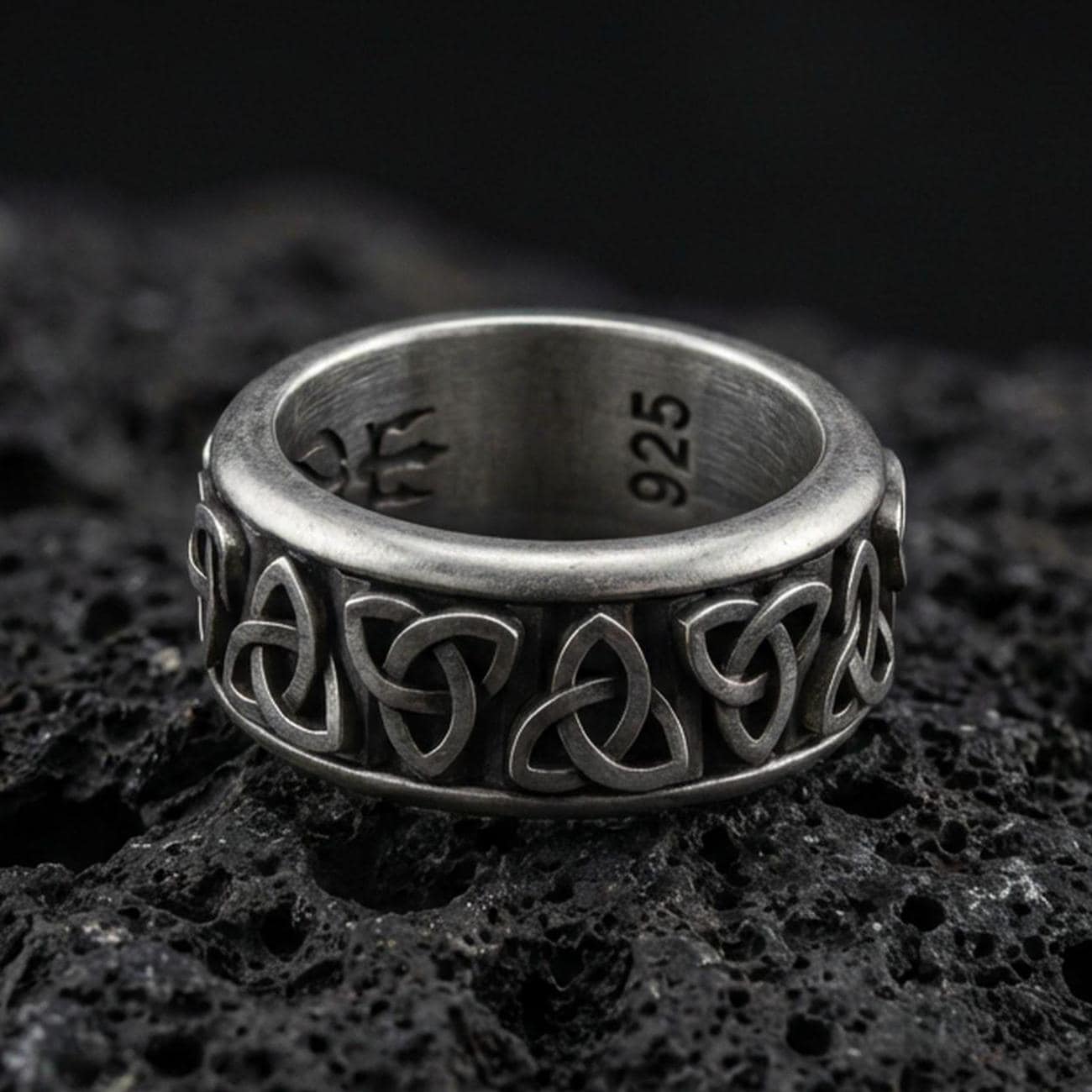 Oxidized Celtic triquetra band ring on textured volcanic rock, centered close-up highlighting the repeating Trinity knot pattern and deep antique shadowed contrast.