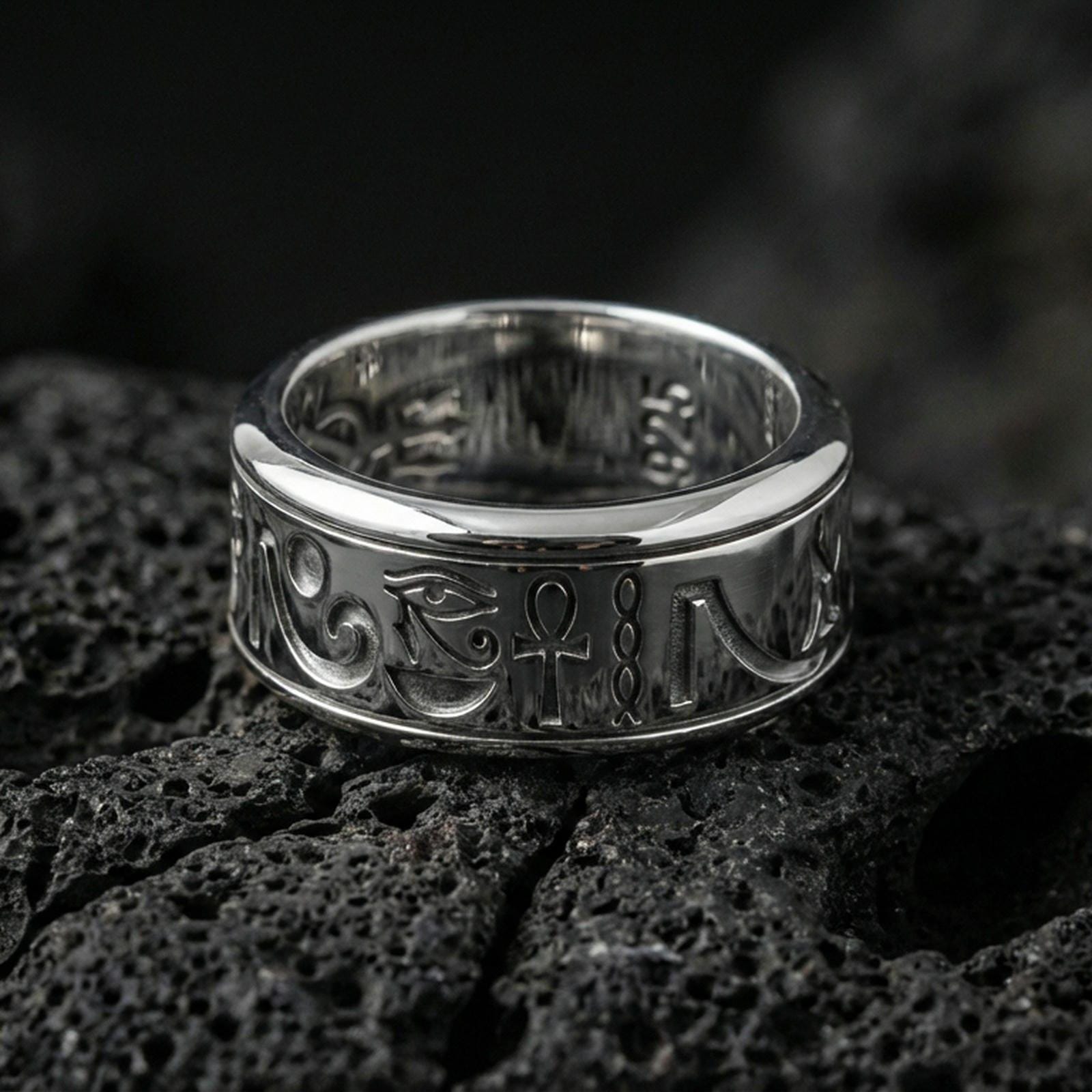 Polished silver Egyptian symbols band ring on textured volcanic rock, crisp macro product photo emphasizing clean edges, sharp hieroglyph engraving, and bright reflective highlights across the band.