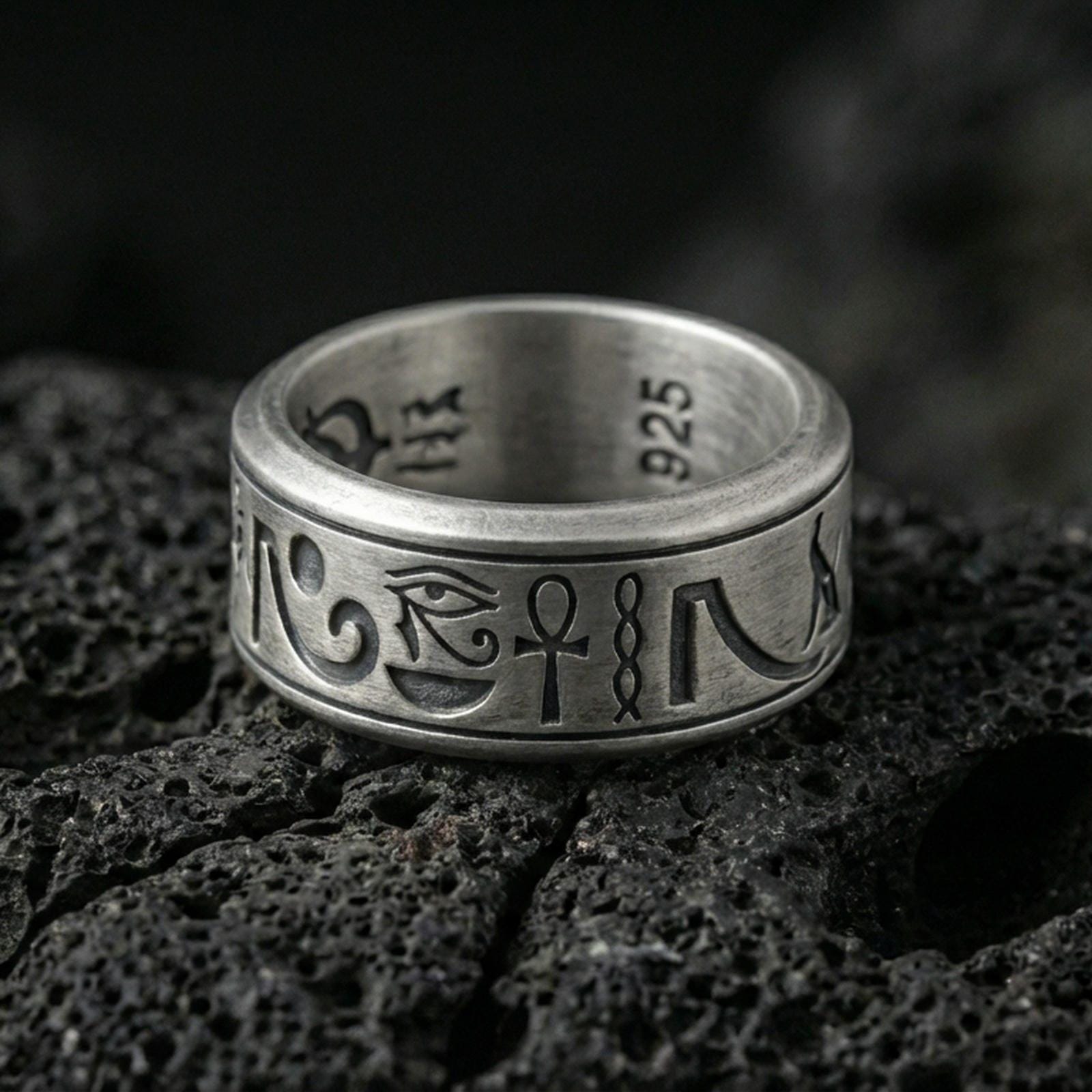 Oxidized Egyptian symbols band ring on textured volcanic rock, centered close-up highlighting the Eye of Horus, ankh motif, engraved glyphs, and deep antique shadowed contrast.