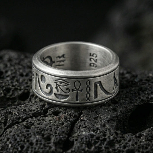 Oxidized Egyptian symbols band ring on textured volcanic rock, centered close-up highlighting the Eye of Horus, ankh motif, engraved glyphs, and deep antique shadowed contrast.