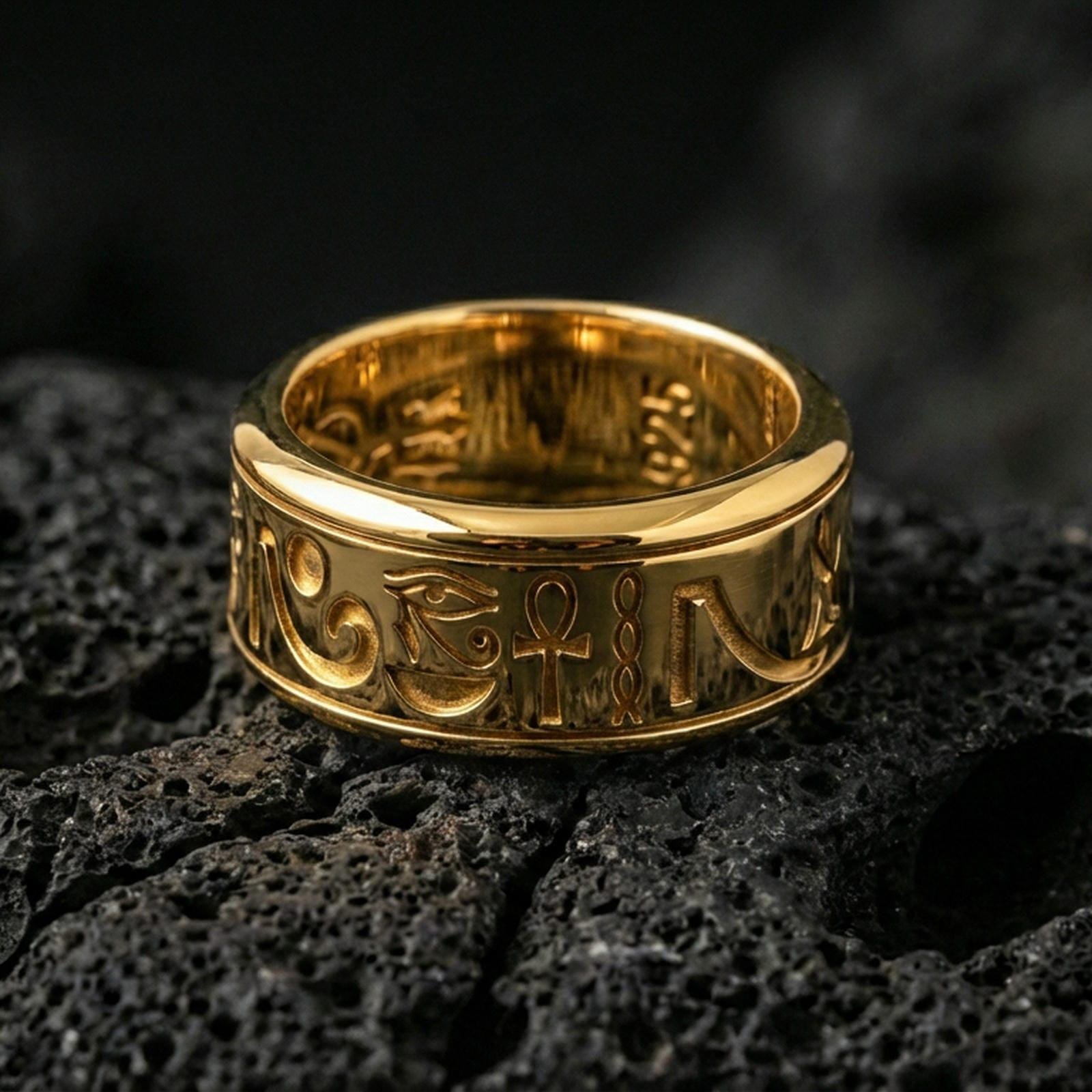 Gold plated Egyptian symbols band ring on textured volcanic rock, premium close-up with warm reflective shine across the Eye of Horus, ankh symbol, and raised engraved lines.