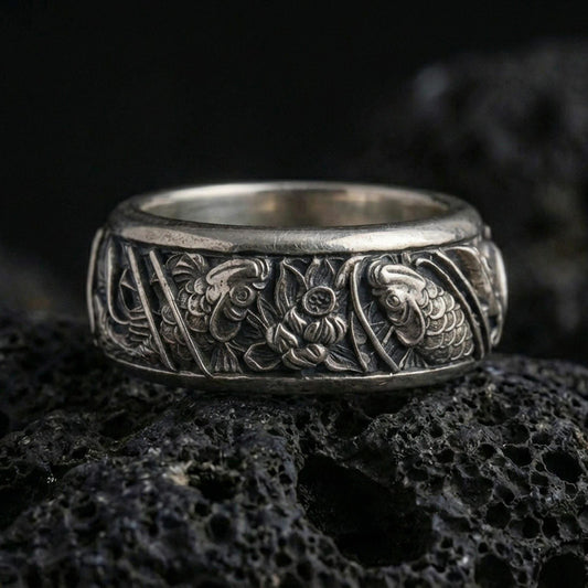 Oxidized koi fish and lotus band ring on textured volcanic rock, centered close-up highlighting the twin koi relief, lotus flower centerpiece, and deep antique shadowed contrast.