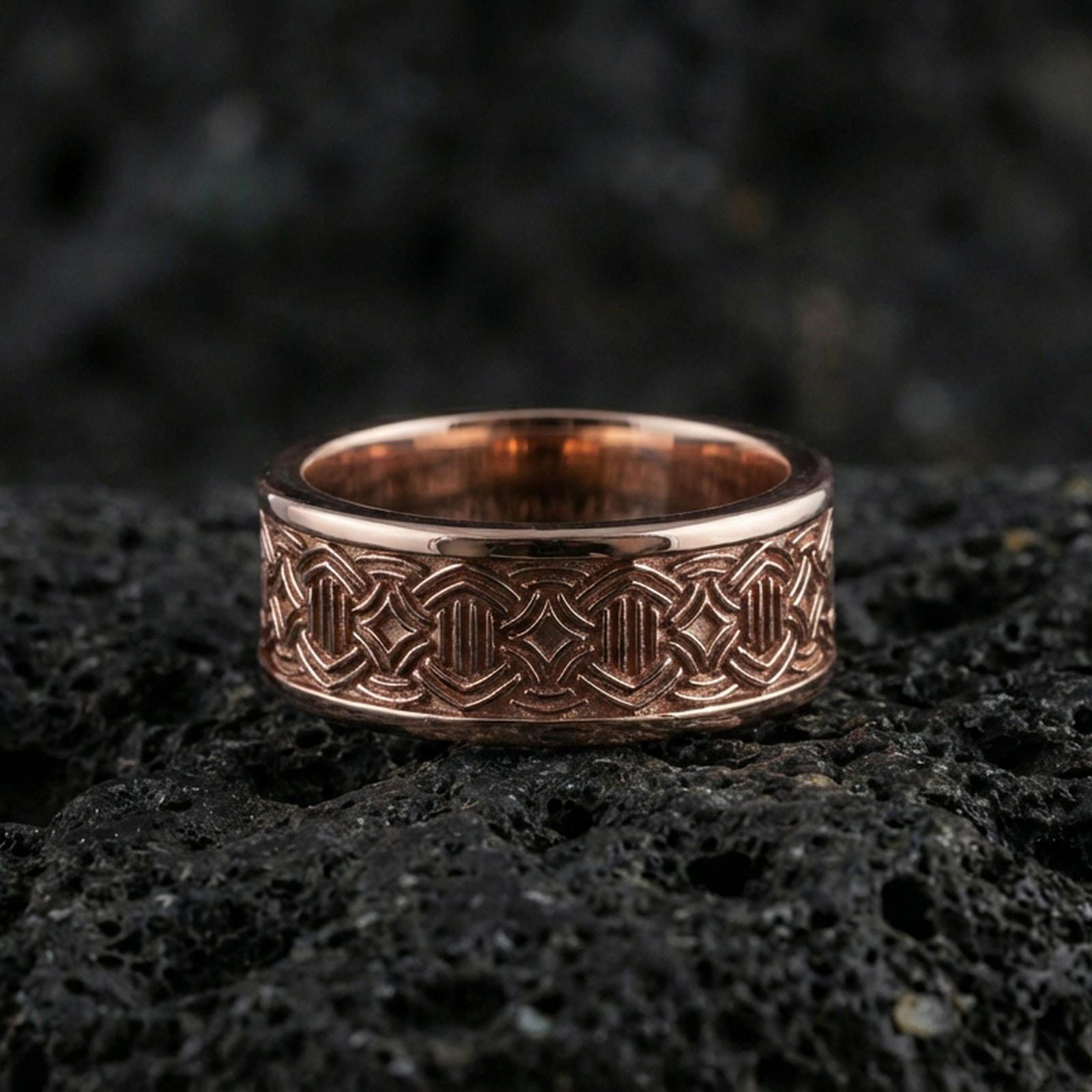 Rose gold plated geometric knotwork band ring on textured volcanic rock, centered studio-style close-up showcasing soft rosy glow and crisp knotwork detailing with a refined finish.