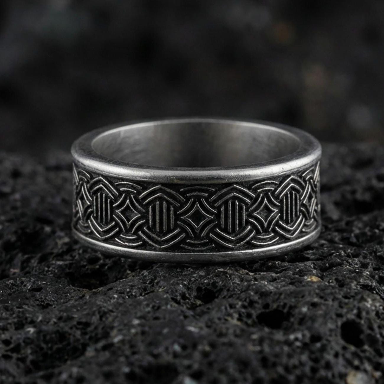 Oxidized geometric knotwork band ring on textured volcanic rock, centered close-up highlighting the repeating interlace pattern, carved depth, and deep antique shadowed contrast.