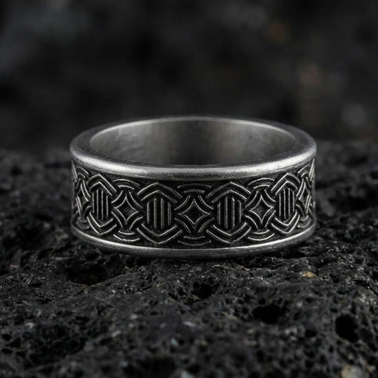 Oxidized geometric knotwork band ring on textured volcanic rock, centered close-up highlighting the repeating interlace pattern, carved depth, and deep antique shadowed contrast.