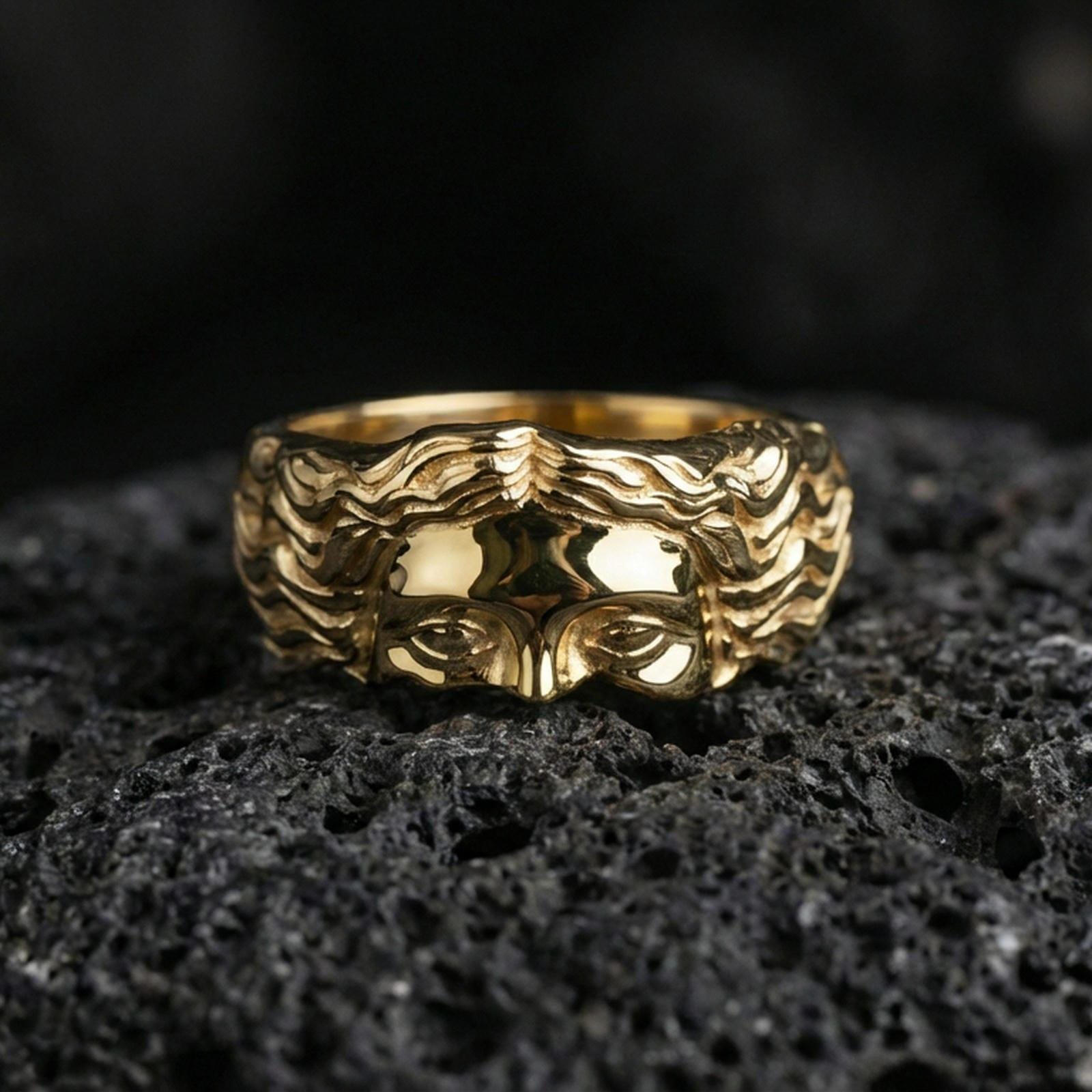 Gold plated face relief band ring on textured volcanic rock, premium close-up with warm reflective shine across the raised face detail and hair texture.