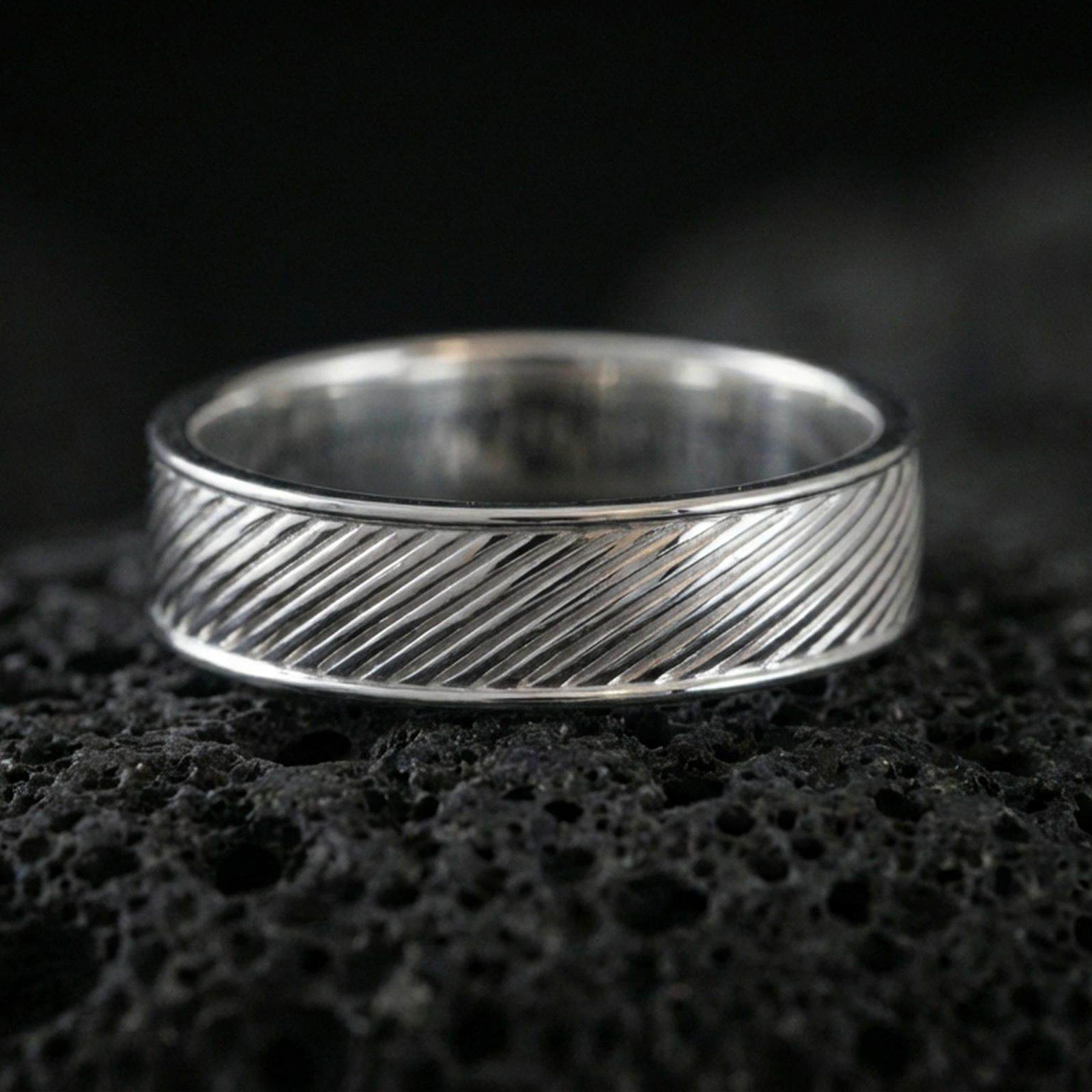 Polished silver diagonal groove band ring on textured volcanic rock, crisp macro product photo emphasizing clean edges, sharp diagonal lines, and bright reflective highlights across the band.
