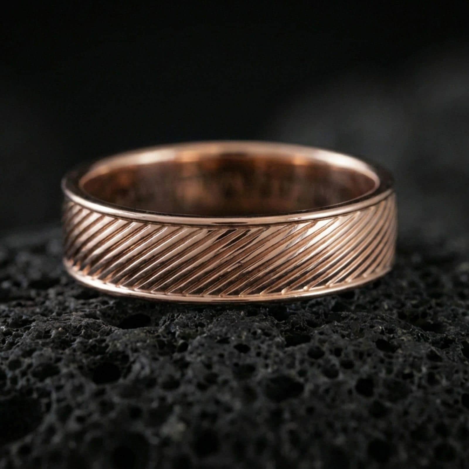 Rose gold plated diagonal groove band ring on textured volcanic rock, centered studio-style close-up showcasing soft rosy glow and crisp diagonal line detailing with a refined finish.