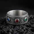 Load image into Gallery viewer, Oxidized ornate gemstone band ring on textured volcanic rock, macro close-up highlighting the antique silver detailing with a deep blue stone, a rich red stone, and a vivid green stone set across the front.
