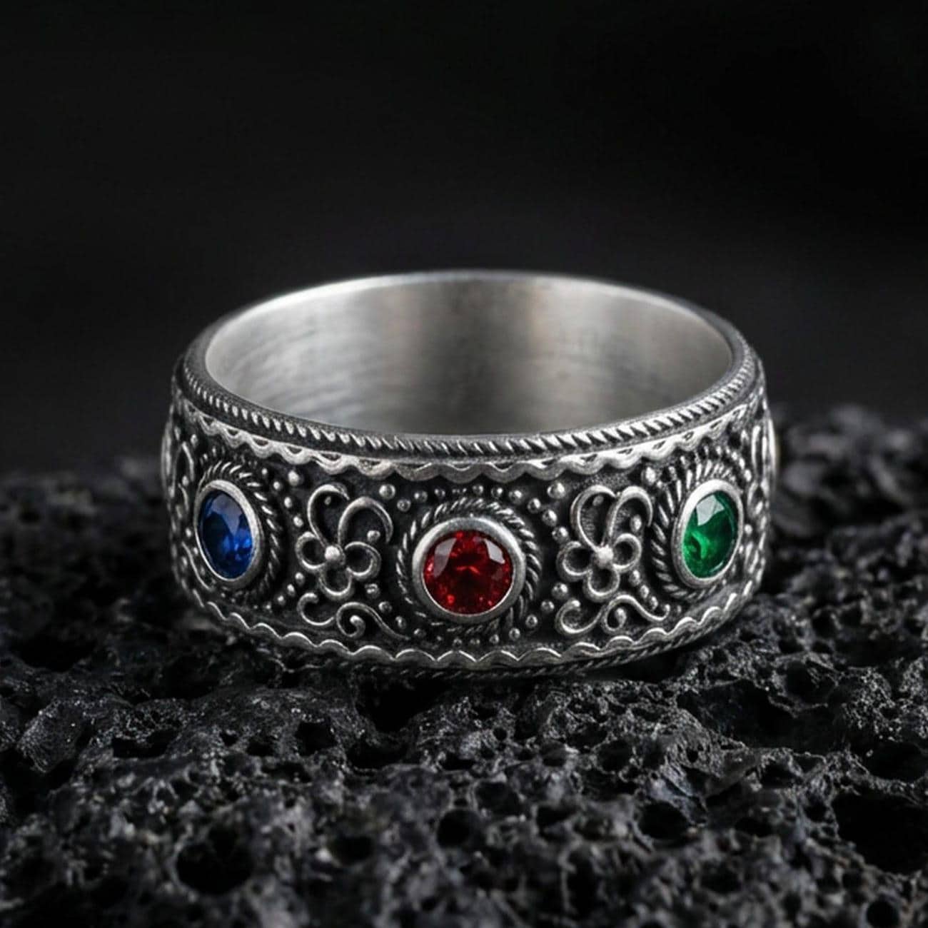 Oxidized ornate gemstone band ring on textured volcanic rock, macro close-up highlighting the antique silver detailing with a deep blue stone, a rich red stone, and a vivid green stone set across the front.