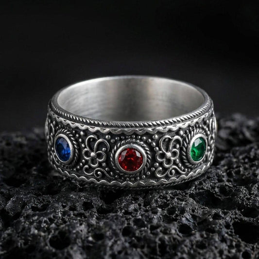 Oxidized ornate gemstone band ring on textured volcanic rock, macro close-up highlighting the antique silver detailing with a deep blue stone, a rich red stone, and a vivid green stone set across the front.