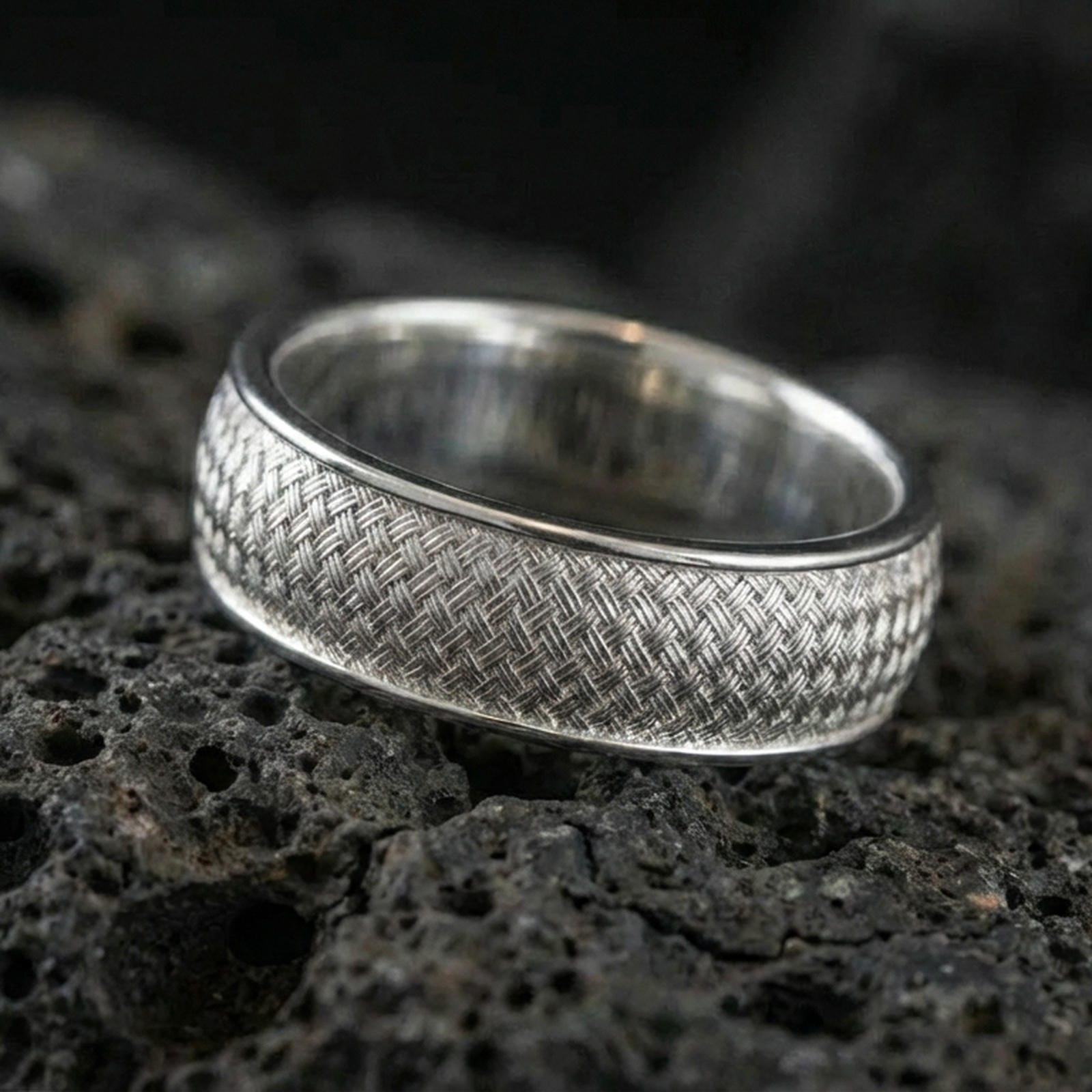 Polished silver woven texture band ring on textured volcanic rock, crisp studio macro emphasizing the basket-weave detail with clean bright reflections.