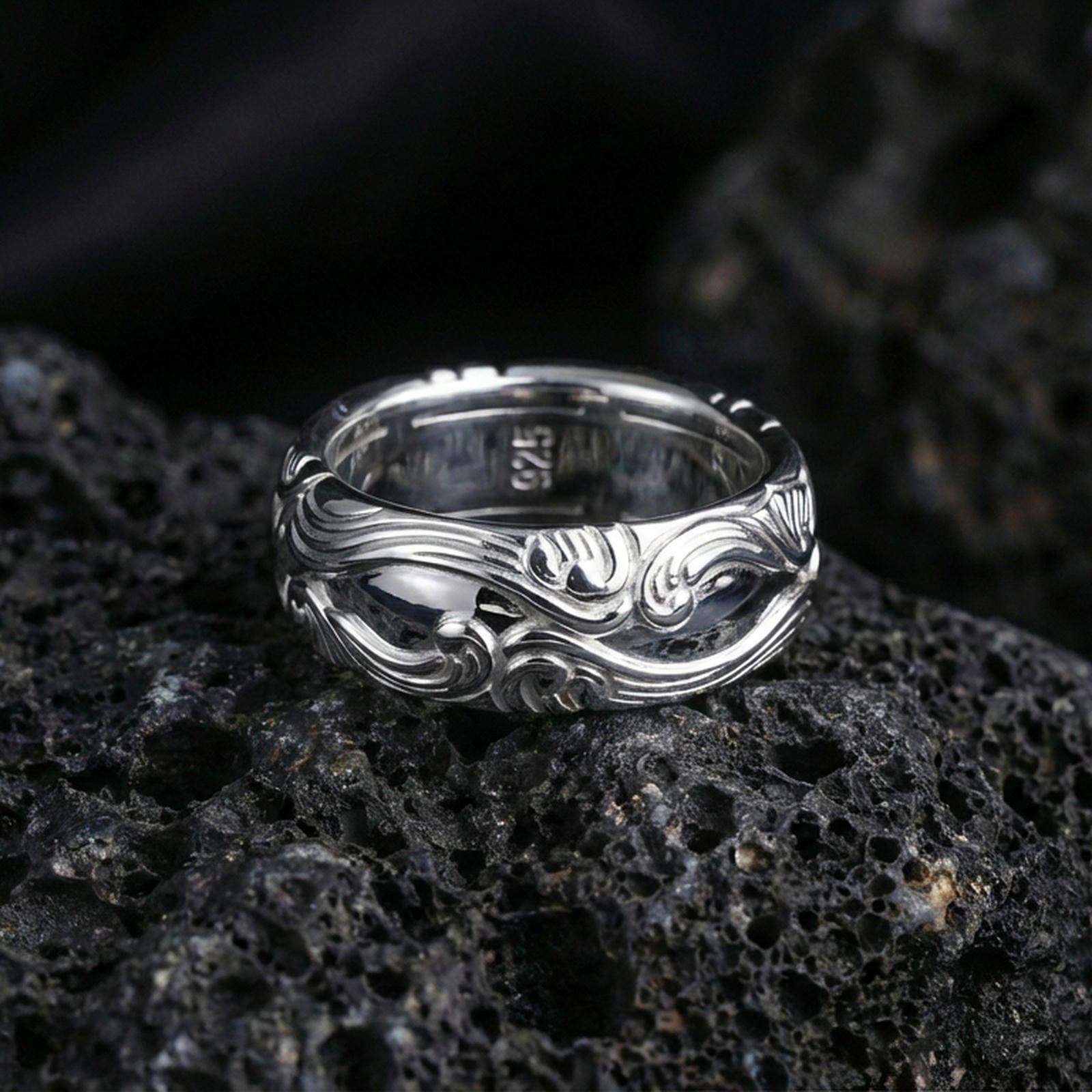 Polished silver scrollwork band ring on textured volcanic rock, crisp studio macro emphasizing clean reflective edges and the carved swirl relief.
