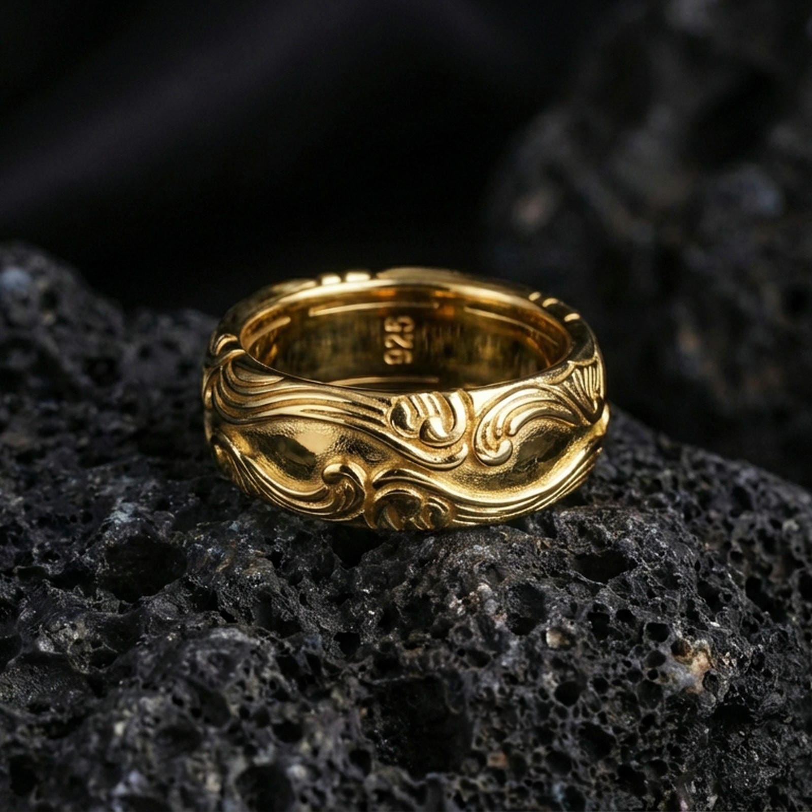 Gold plated scrollwork band ring on textured volcanic rock, premium close-up showing warm gold tone across the raised wave patterns with smooth rounded profile.