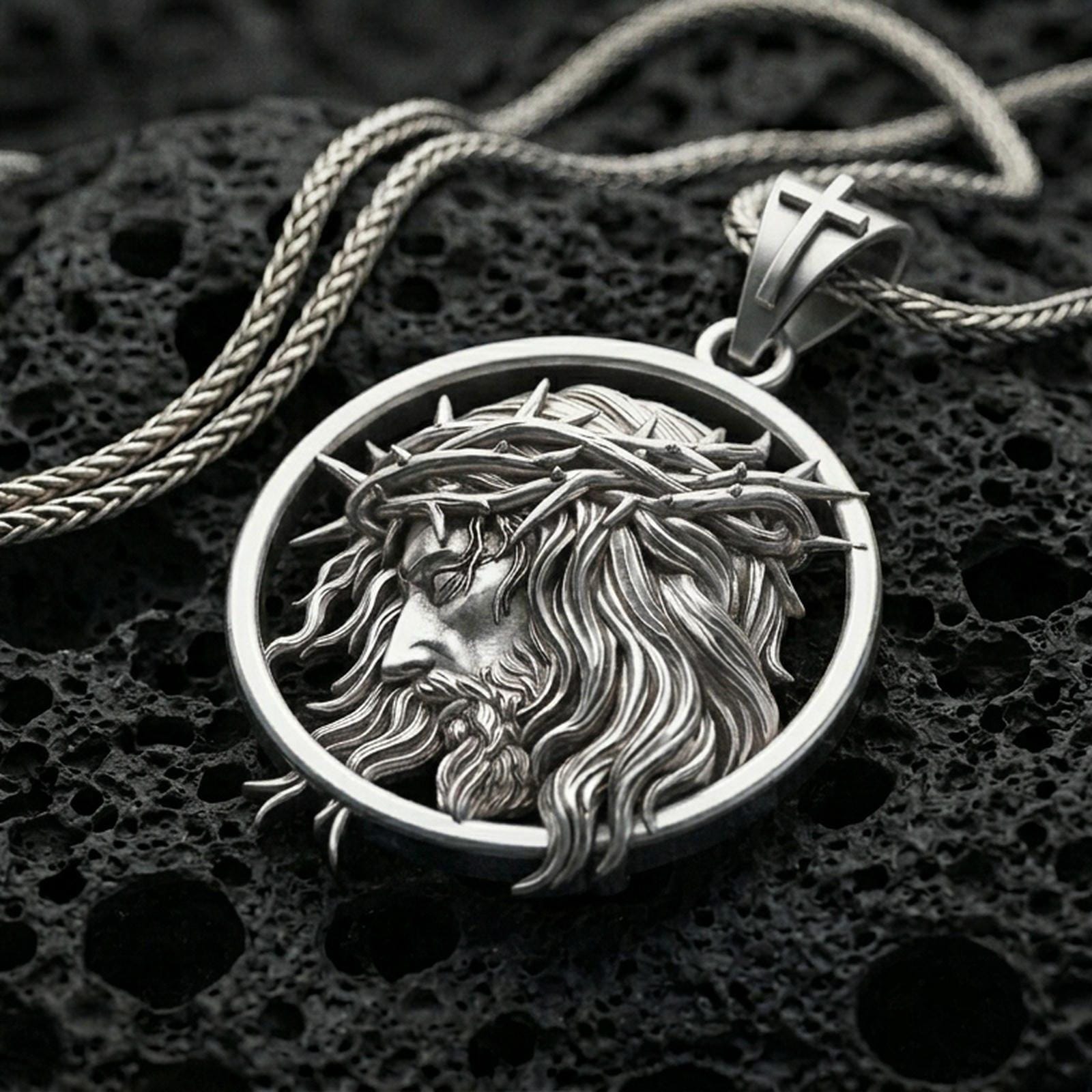 Polished silver Jesus medallion with circular frame, crown of thorns, and sculpted side profile placed on textured volcanic rock in a premium studio product shot