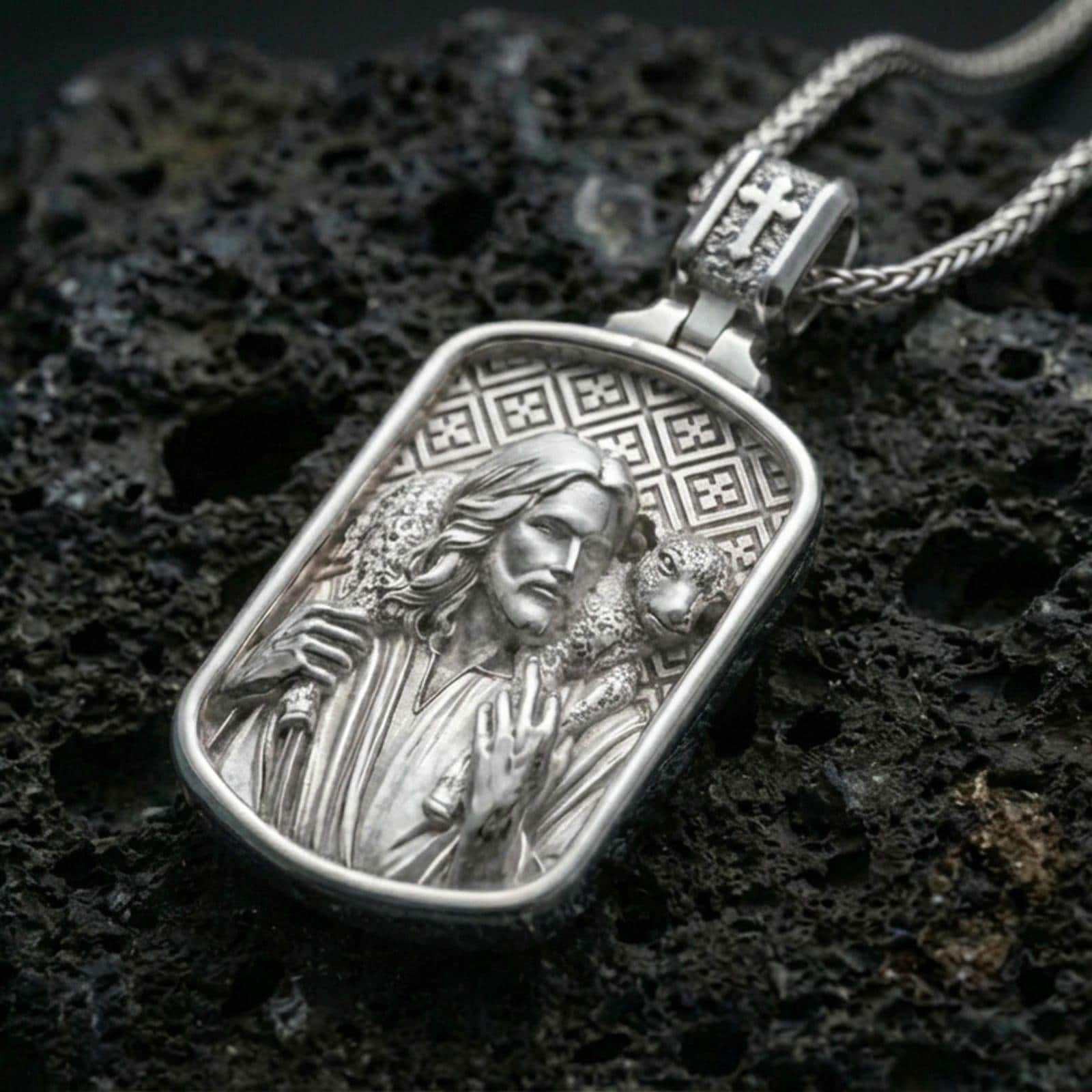 Polished silver Jesus medallion with shepherd motif, lamb detail, and rounded rectangular frame placed on textured volcanic rock in a premium studio product shot