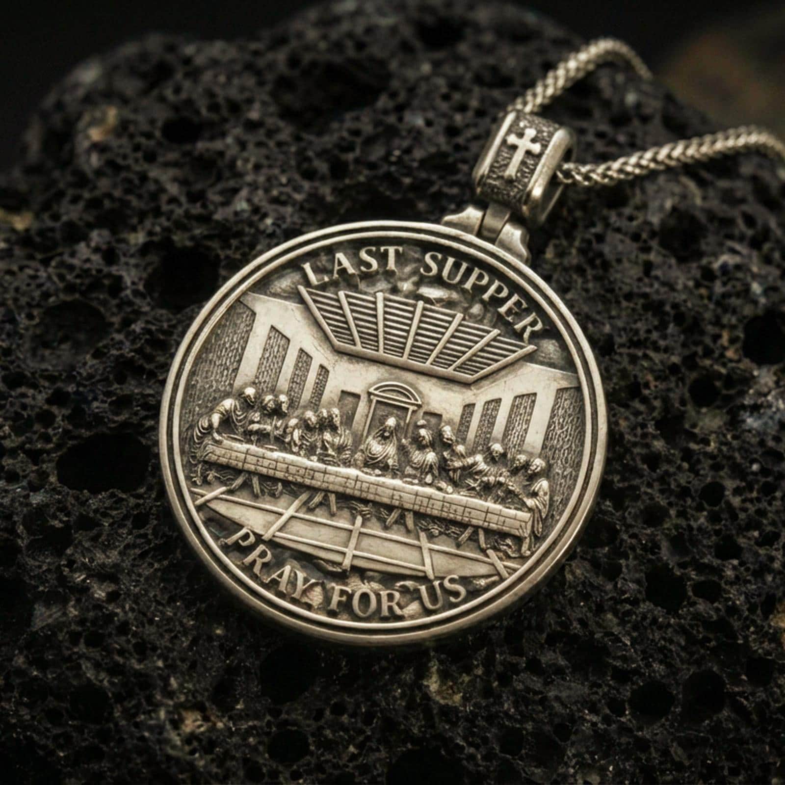 Bronze Last Supper pendant with Pray for Us inscription, detailed round medallion design, and two-sided devotional concept displayed on dark volcanic rock in a close-up luxury jewelry composition
