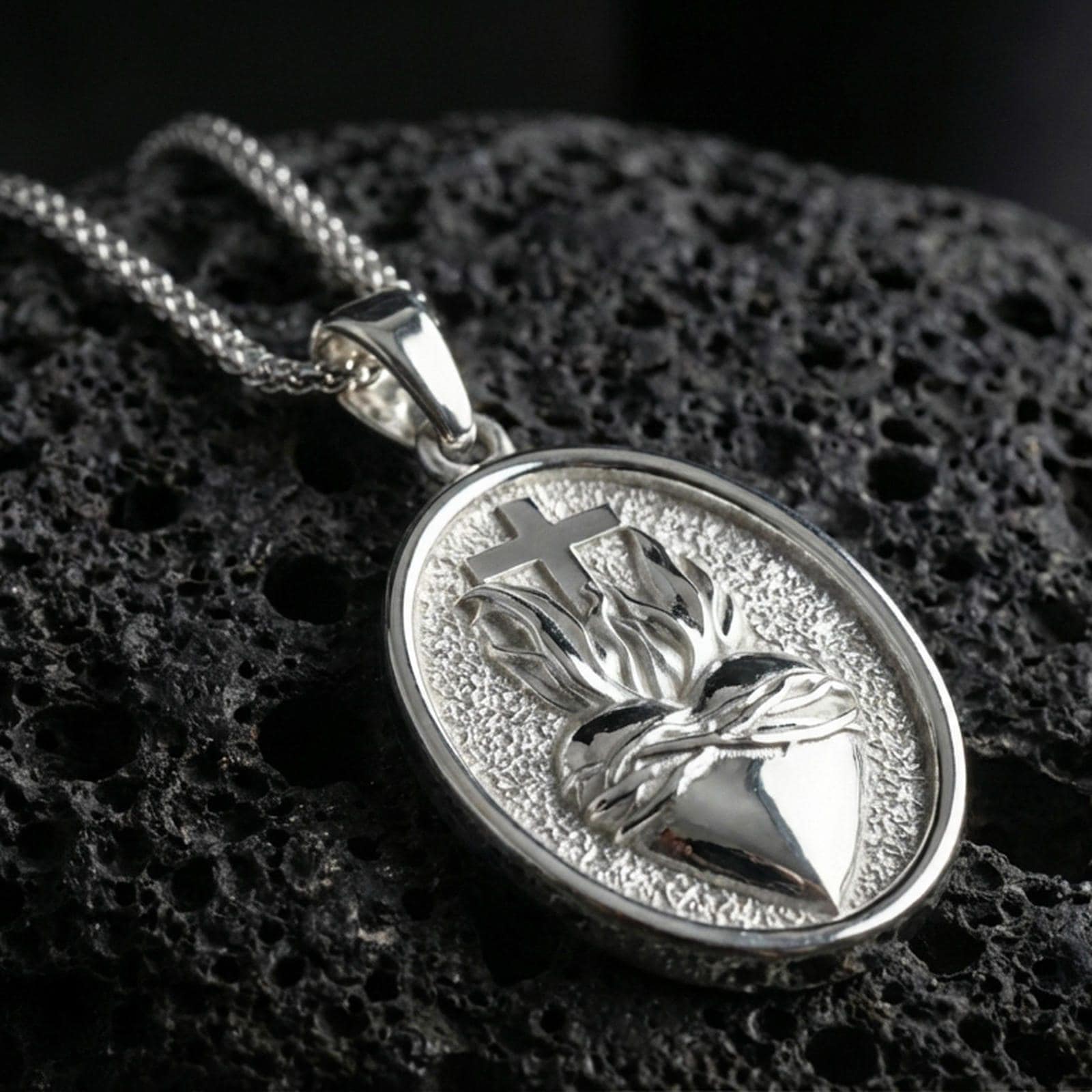 Polished silver Christian medal with Sacred Heart motif, radiant flame relief, and smooth oval frame placed on textured volcanic rock in a premium studio product shot