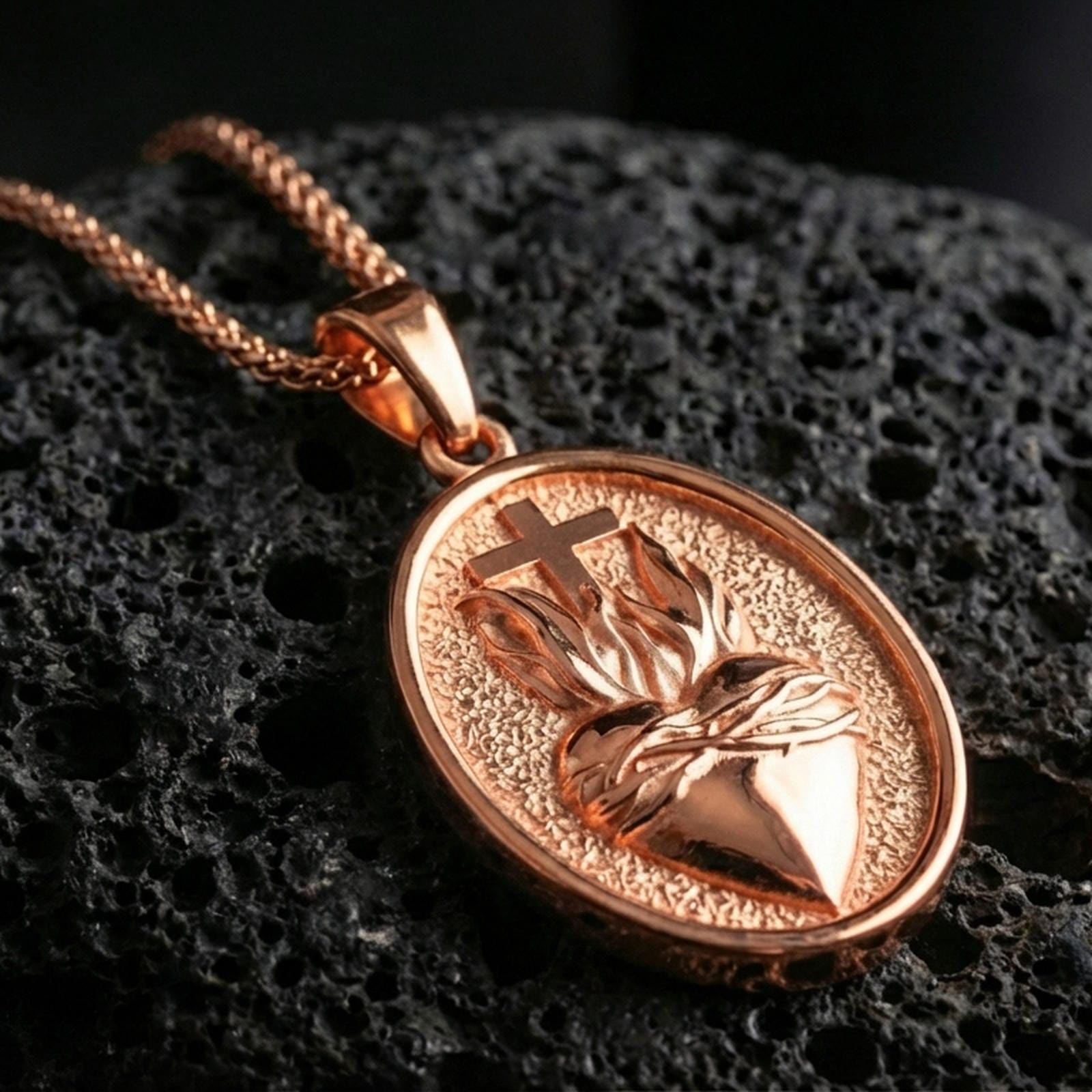 Rose gold plated oval Christian pendant with Sacred Heart relief, flame accent, and classic devotional silhouette resting on volcanic rock in an elegant close-up product presentation