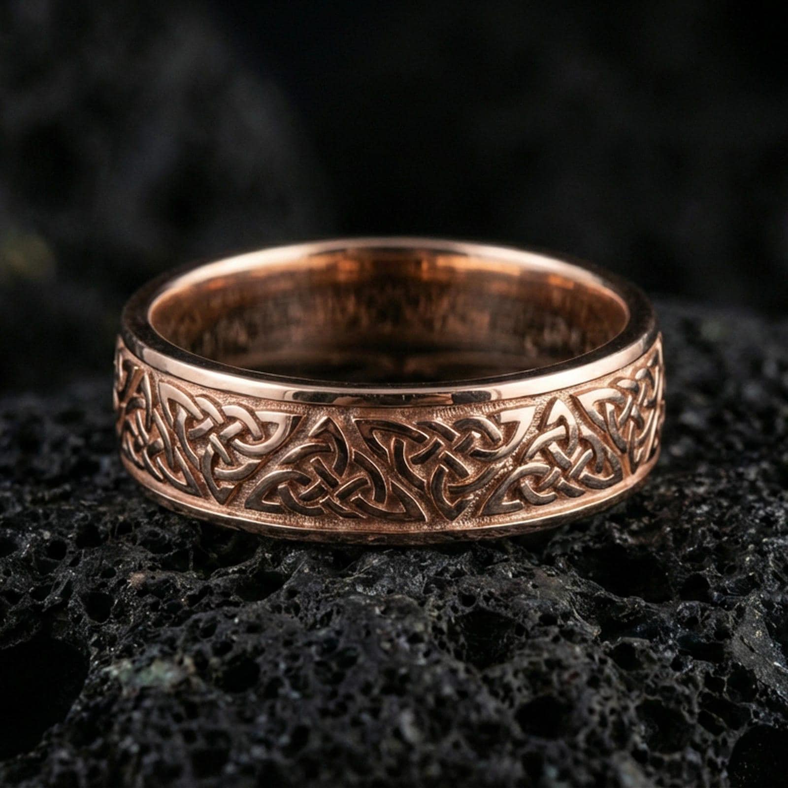 Rose gold plated Celtic knot band ring on volcanic stone, refined close-up with soft rosy highlights and clear interwoven Celtic knot texture.
