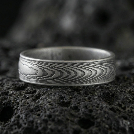 Oxidized Damascus-style band ring on volcanic stone, macro close-up highlighting darkened layered wave lines and brushed edges.