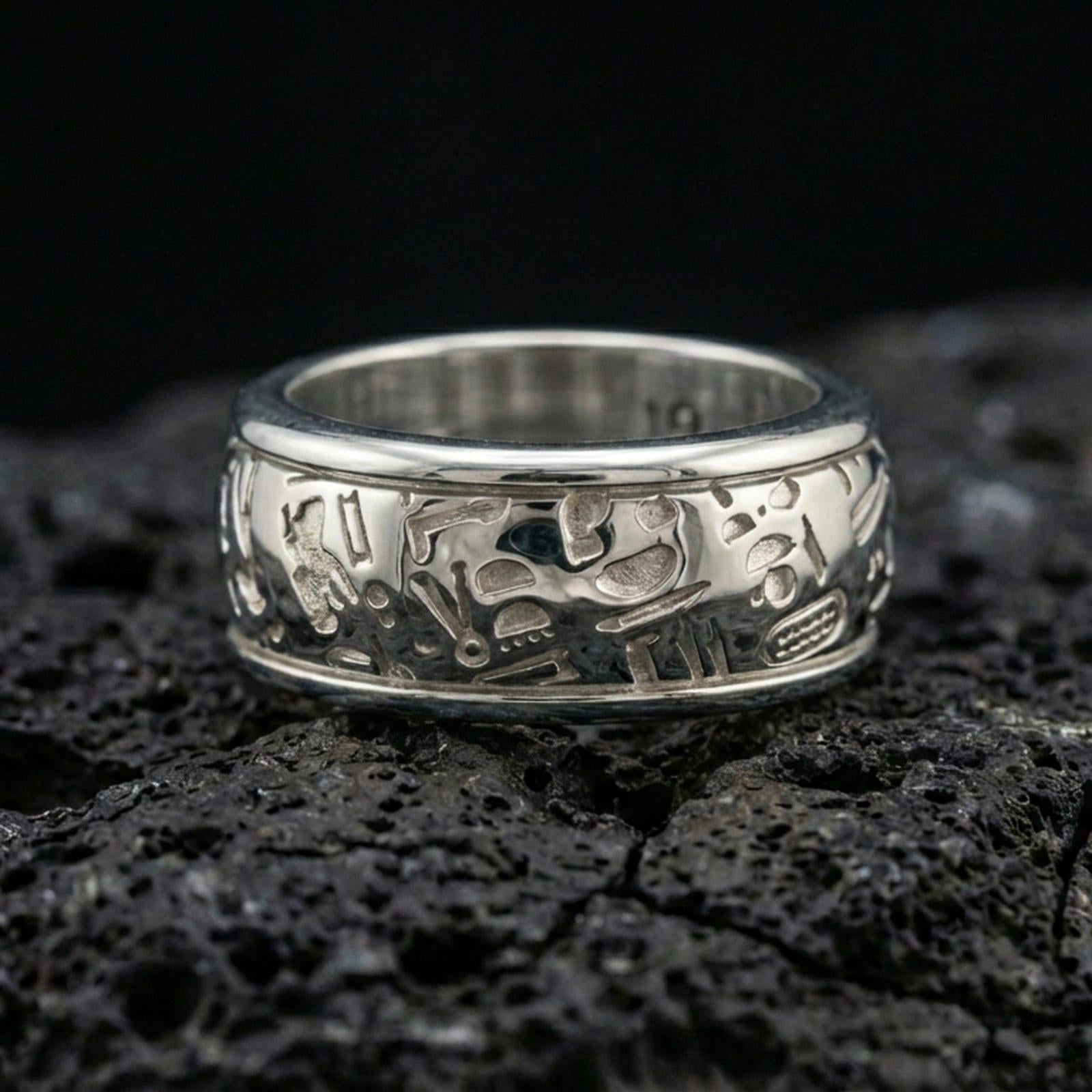 Polished silver hieroglyph band ring on volcanic stone, crisp macro product photo emphasizing clean engraved glyph details and bright reflective edges.