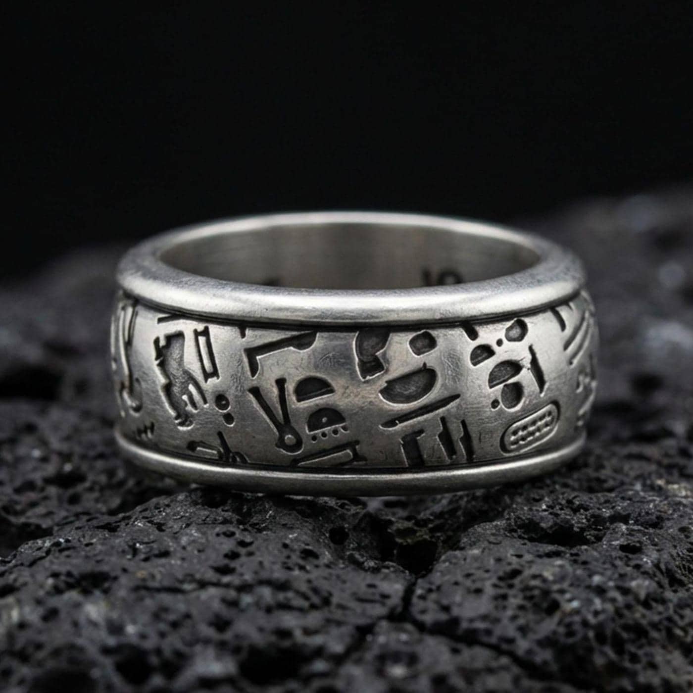 Oxidized sterling silver hieroglyph band ring on volcanic stone, macro close-up highlighting deep-cut Egyptian symbols and matte patina texture.