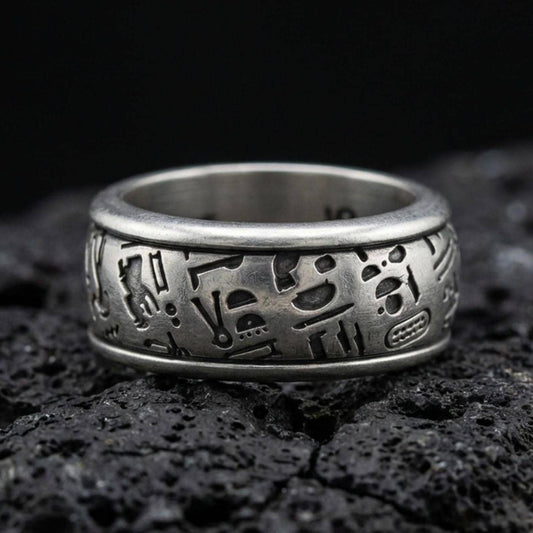 Oxidized sterling silver hieroglyph band ring on volcanic stone, macro close-up highlighting deep-cut Egyptian symbols and matte patina texture.