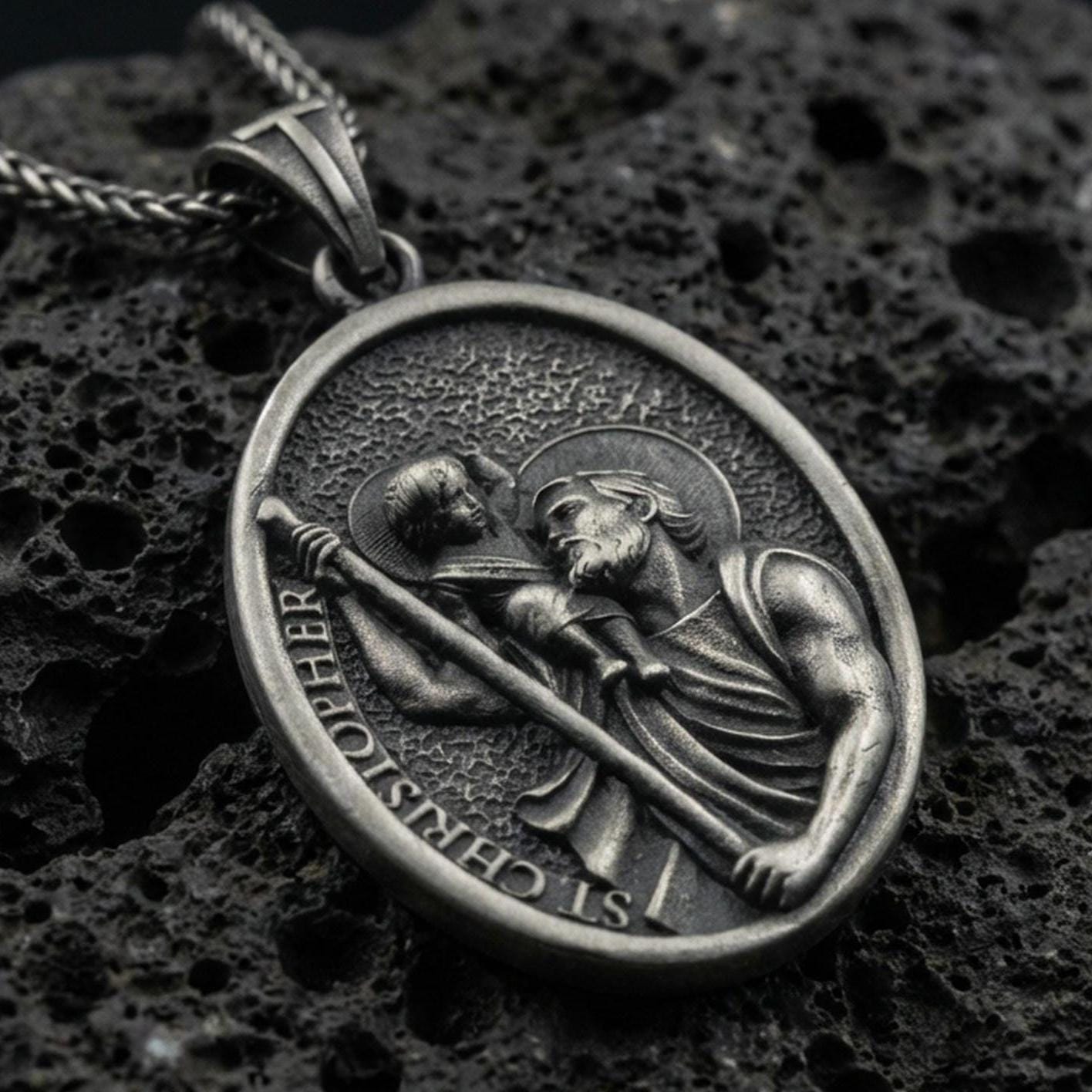 Oxidized Saint Christopher pendant with Child Jesus, staff detail, and oval devotional portrait displayed on dark volcanic rock in a close-up luxury jewelry composition
