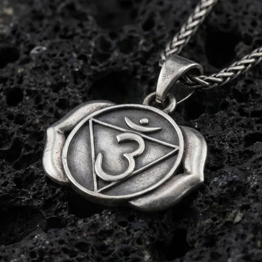 Oxidized third eye chakra pendant with Om symbol, triangle motif, and two-petal lotus design displayed on dark volcanic rock in a close-up luxury jewelry composition