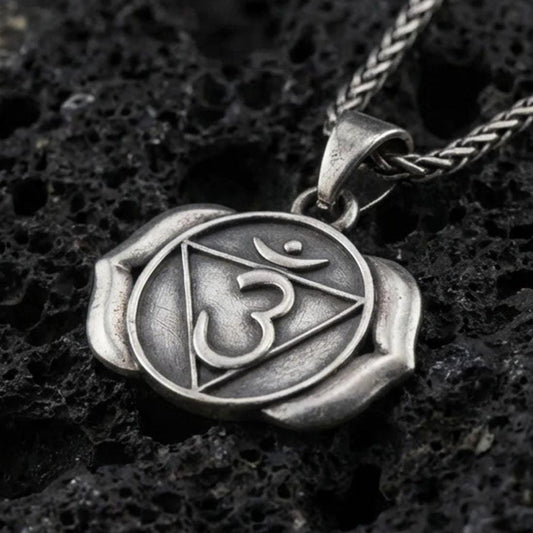 Oxidized third eye chakra pendant with Om symbol, triangle motif, and two-petal lotus design displayed on dark volcanic rock in a close-up luxury jewelry composition