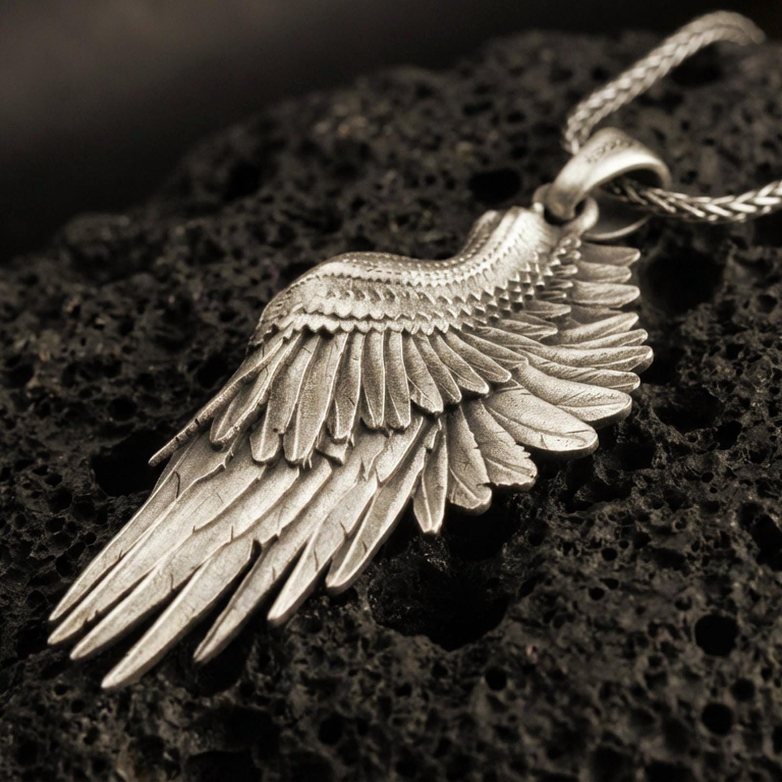 Bronze angel wing pendant with layered feather detailing and elongated wing silhouette displayed on dark volcanic rock in a close-up luxury jewelry composition