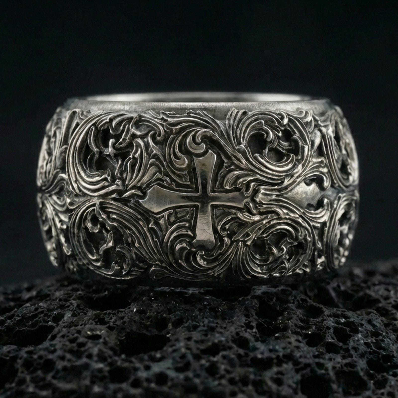 Oxidized sterling silver ornate cross band ring on volcanic stone, macro close-up highlighting deep relief scrollwork and antique contrast around the cross motif.